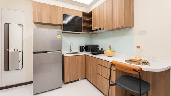 L-shaped apartment kitchen with light wood-look cabinets, a stainless steel refrigerator, white countertops, and a clear glass backsplash.