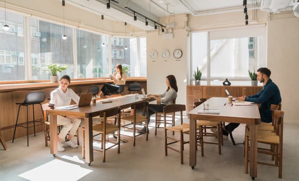 Bright, modern coworking space with large windows overlooking a city view, featuring long communal wooden tables, mid-century style chairs, and several people working on laptops and reading.