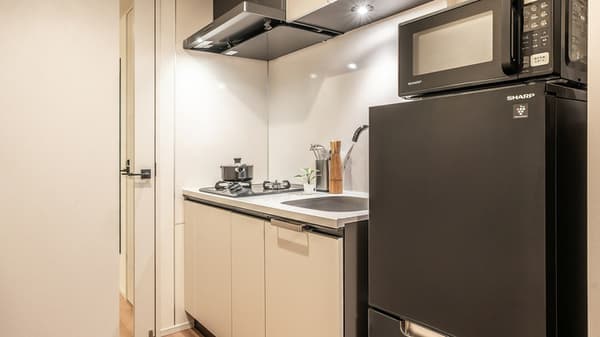 Compact galley kitchen featuring a black refrigerator and microwave, light beige cabinets, a sink, and a two-burner cooktop with a pot and pan.