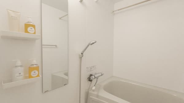 View inside a bright, white Japanese-style bathroom featuring a bathtub, a shower head, and a large mirror with shelves holding toiletries.