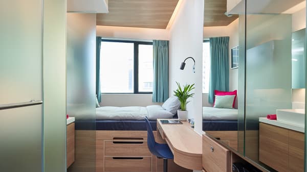 A compact studio apartment featuring a double bed with under-bed storage drawers, a light wooden desk, and large mirrors and frosted glass partitions that reflect the space and window.