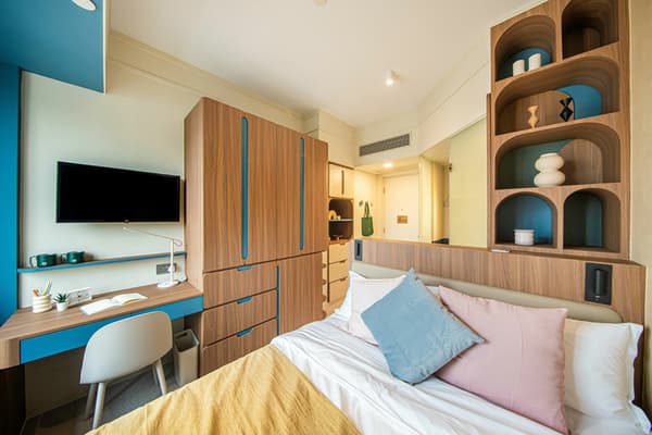 View of a modern, compact bedroom space with light walls and colorful accents. The room features a bed with pastel pillows, a built-in desk with a white chair and a wall-mounted TV, and floor-to-ceiling wood cabinetry with drawers for storage. A tall, decorative shelving unit with curved recesses stands to the right.