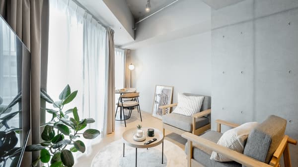 Modern living space with two light wood and gray armchairs, a small round coffee table, a tall window with light curtains, and an exposed concrete accent column.