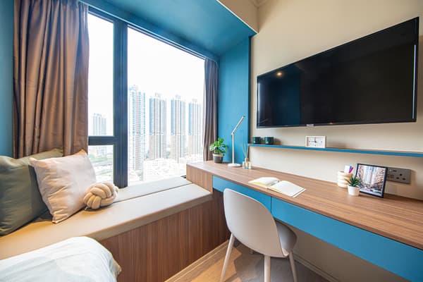 A bright modern desk area next to a large floor-to-ceiling window with brown curtains. The desk has a wood top with a blue accent panel, a chair, and a wall-mounted TV. Below the window is a cushioned window seat with a city skyline view of tall buildings.