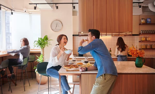 A group of people sharing a meal in a bright, modern communal kitchen and dining area, with a smiling couple in the foreground eating pizza and salad at a marble-top island counter.