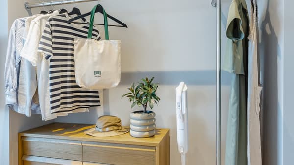 Close-up of an open wardrobe area showing clothes hanging on a rail, a wooden dresser below with decor, and a white, upright vacuum cleaner beside it.