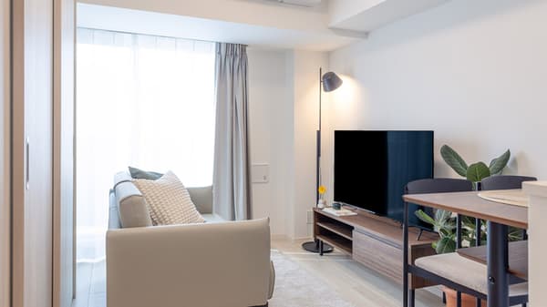View of the living room featuring a light gray sofa, a floor lamp, a large TV on a wooden console, and sheer curtains covering a window.