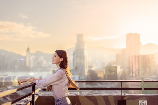 A woman in a white shirt and jeans stands on a rooftop deck, smiling as she looks out over the wide, sunlit Hong Kong cityscape and mountains at sunset.