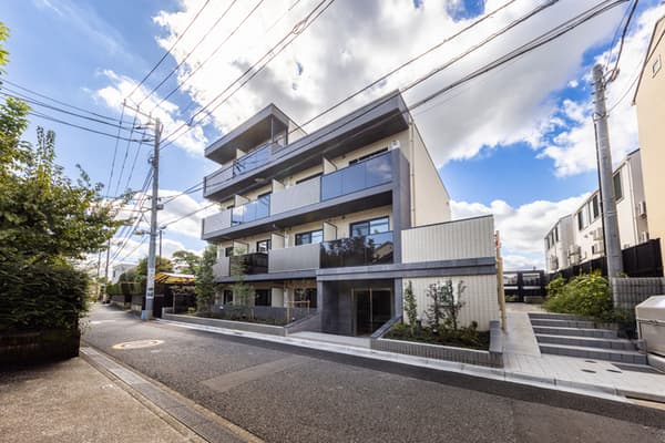 Exterior street-level view of a modern, three-story apartment building with dark grey and light beige accents, glass balcony railings, and simple landscaping under a blue, cloudy sky.