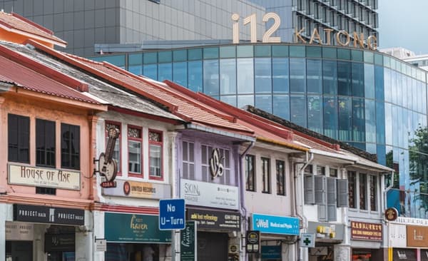 A row of colorful, traditional shophouses with small businesses, contrasting sharply with the modern, glass-facade i12 Katong mall towering behind them.