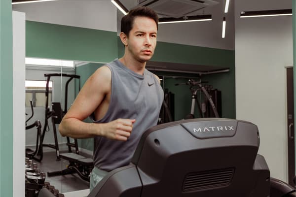 Close-up shot of a man wearing a gray tank top running on a Matrix brand treadmill in a well-equipped gym with green and white walls and mirrors.