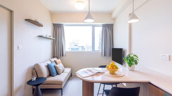 Bright studio living space with a beige sofa, a round dining table/bar with stools, and white pendant lighting, all centered around a window.