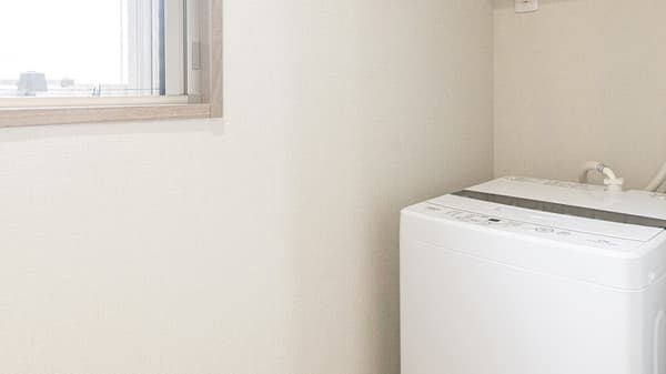 Utility area featuring a white top-loading washing machine beneath a small window, with light beige textured wallpaper on the walls in Monzennakacho.