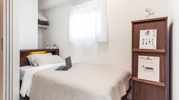 A bright, minimalist single bedroom with a wooden bed frame, back-lit headboard, laptop, and a tall, narrow wooden shelving unit.
