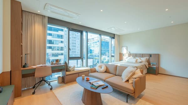 Wide interior shot of the spacious Junior Luxe Suite, featuring a queen-size bed, a tan leather two-seater sofa lounge area, a dedicated window-facing desk, and floor-to-ceiling windows with city views.