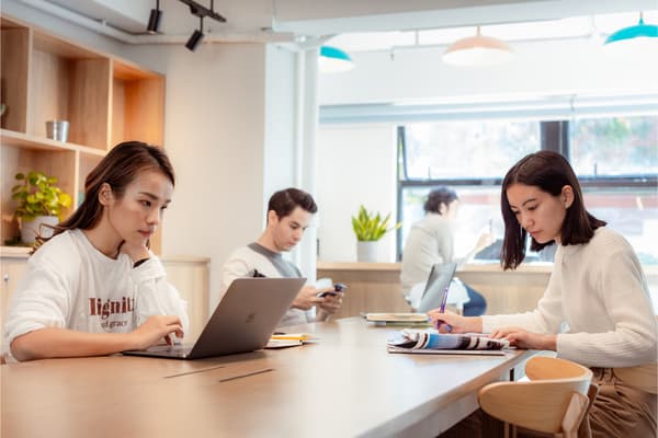 Diverse group of young professionals working at a long communal table in a bright co-working space, using laptops and writing in notebooks with wooden shelves visible in the background.