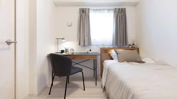 Second bedroom featuring a single bed with light wood headboard and a small study area with a wooden desk, gray chair, and desk lamp next to a curtained window.