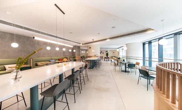 An expansive, bright lounge area with various seating options, including a long white communal table with bar stools, green banquette seating along a textured wall, and smaller round tables near the large windows on the right.