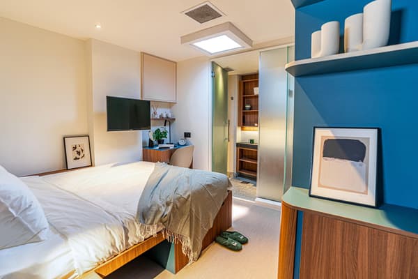 Compact, modern bedroom featuring a queen bed, a skylight, a small desk area, a mounted TV, and striking teal blue wall accents with built-in shelving.