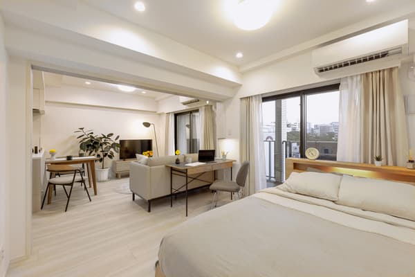 A wide-angle view of a bright, open-plan studio apartment showing the bedroom in the foreground, a dedicated work desk area, and a living/dining area visible through a wide doorway.