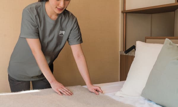 Professional housekeeping staff member wearing a grey uniform neatly making the bed in a modern apartment bedroom.