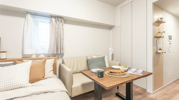 Closer view of the studio's living space, showing a light gray sofa, wooden dining table, and built-in white cabinets with integrated shelving.