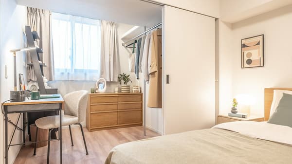 Bright bedroom corner featuring a work desk and chair next to a large window, a light wood dresser, and an open closet area with hanging clothes storage.