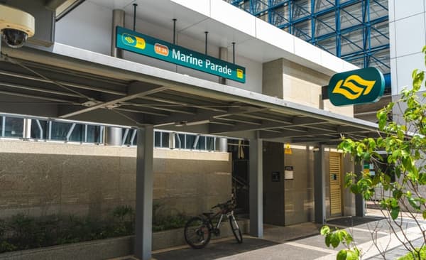 The modern, sheltered entrance of the Marine Parade MRT (TE26) station with its signage visible, and a bicycle parked underneath the canopy.