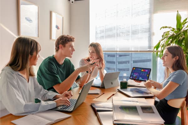 A diverse group of young professionals collaborating around a table in a bright, modern meeting room with laptops and a tablet displaying data.