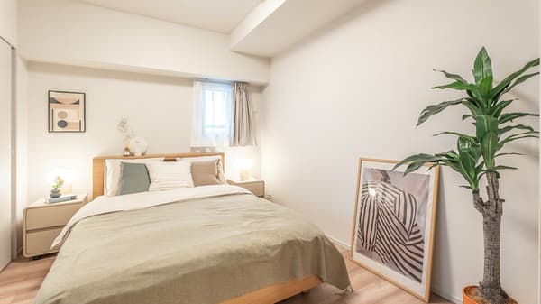Light-filled bedroom with a wooden double bed, neutral-toned bedding, two small bedside tables with lamps, and a large framed artwork next to a tall potted plant.