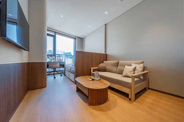 Wide view of a modern studio apartment with a queen bed and a separated sofa bed lounge area featuring a unique adjustable height coffee table, set near a large window.