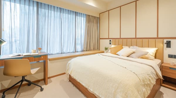 Modern junior suite bedroom with a queen bed, upholstered headboard, dedicated desk area, and a large window with sheer curtains and a cushioned seat.