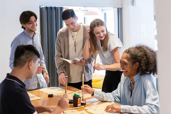 A group of smiling young adults enjoying a social activity, learning or practicing Chinese calligraphy together at a wooden table in a communal space.