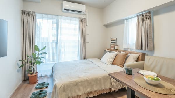 Compact studio apartment with a double bed, small two-seater sofa, and a low dining table, illuminated by natural light from a large window.