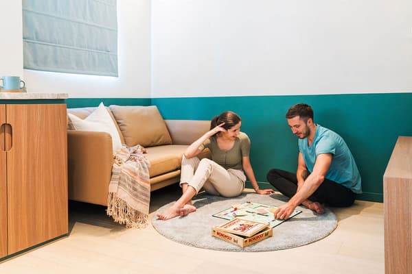 A young couple sitting on a light gray round rug on the floor, playing a game of Monopoly in a modern living area with a neutral-colored sofa, a blue-green accent wall, and light wood flooring.
