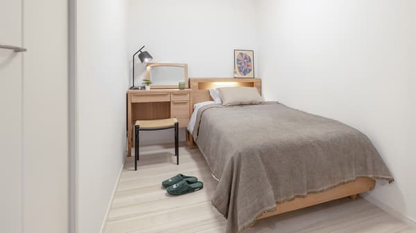 Compact second bedroom with a single bed, light wood desk/vanity, and a woven stool, styled with minimalist wall art and house slippers on the floor.