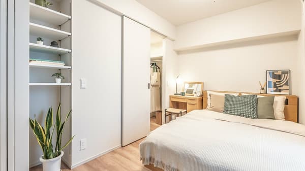 Simple apartment bedroom with natural wood bed frame, side table, a small desk area, and a built-in shelf unit, with sliding doors leading to a closet.