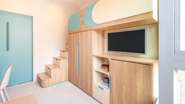 Interior view of a studio apartment showing a multi-functional wooden storage and bed unit with built-in stairs, a closet, and a mounted TV, next to a pale blue door.