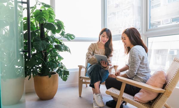 Two women sitting in modern woven-back wooden armchairs by a large window, with one woman showing the other content on a digital tablet next to a large potted monstera plant.