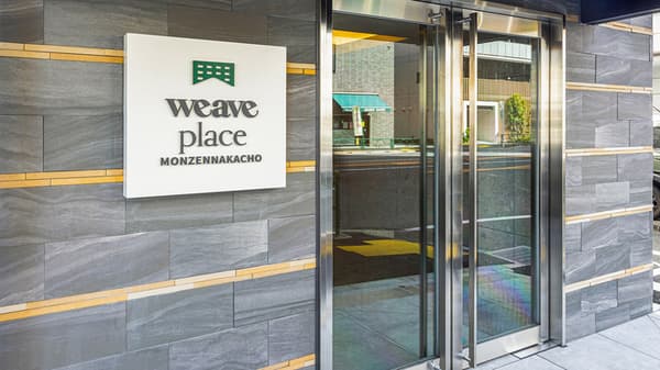 Exterior view of the main entrance to Weave Place Monzennakacho, featuring a contemporary grey slate wall with gold accents, a white sign with the green 'weave place' logo and name, and large glass double doors.