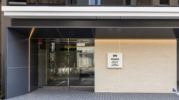 The modern main entrance of Weave Place Asakusa Kaminarimon, featuring a glass door, dark gray paneling, and a light brick wall with the building's logo sign.