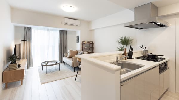 Wide-angle view of a bright, open-concept living room and kitchen, showing a light leather sofa, wood TV console, and kitchen counter with a sink and stovetop.