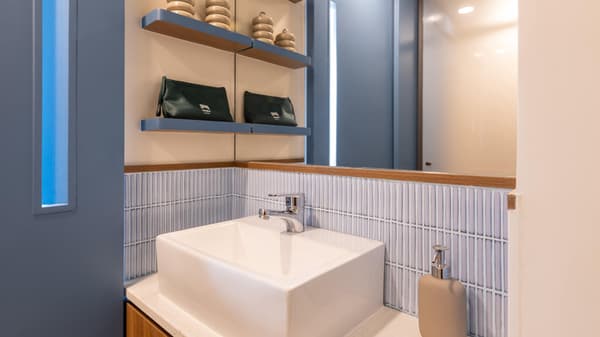Modern bathroom vanity with a square sink, ridged pastel blue tile backsplash, wooden trim, and blue shelves holding small cosmetic bags.