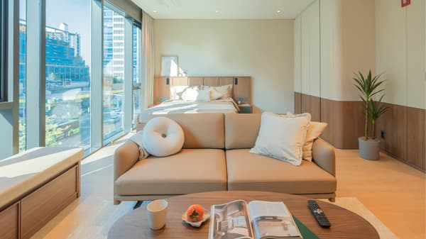A close view of the bright living area in the Junior Luxe Suite, centered around a tan leather sofa and a round wooden coffee table, with a queen bed visible in the background and panoramic windows spanning the wall.
