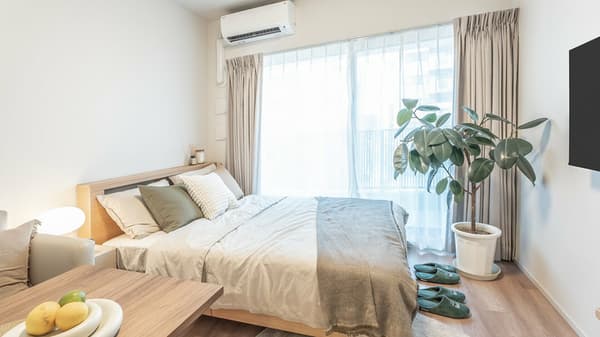 Cozy sleeping area in a 1K apartment with a double bed, light wood frame, neutral bedding, an air conditioner, and a large potted plant next to the balcony window.