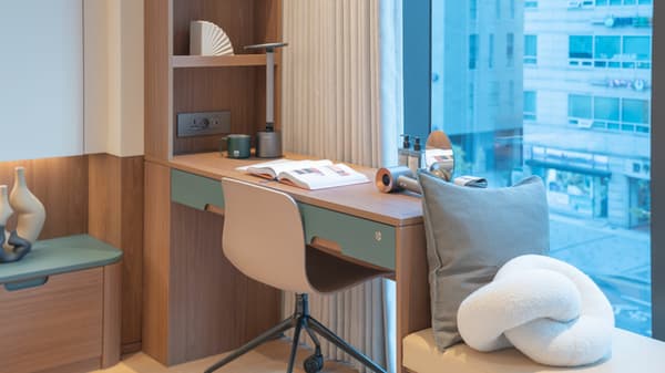 Close-up of the built-in desk workspace with light wood and subtle green drawers, featuring integrated shelving, power outlets, a desk lamp, and a view of the street through the large window.
