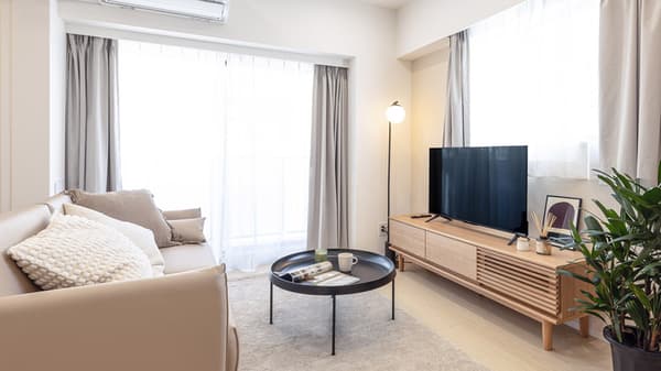 Furnished living room with a neutral-toned sofa, a modern wooden TV stand, a flat-screen television, a round black coffee table, and large windows with sheer and grey curtains.