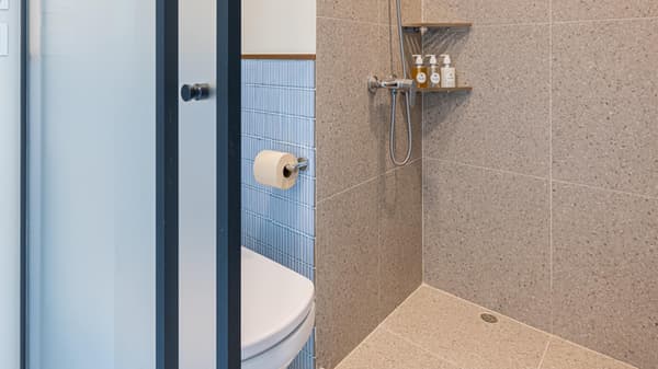 View into the shower area of a compact bathroom, showing large beige speckled terrazzo-style wall and floor tiles next to the toilet area behind a glass partition.
