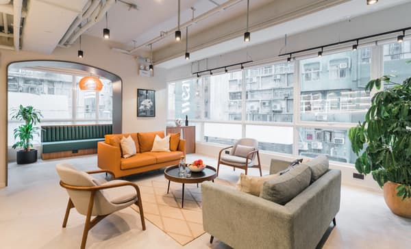 Stylish residential common area lounge with large windows, featuring a bright orange sofa, a gray loveseat, and two mid-century style armchairs arranged on a rug around a low coffee table.