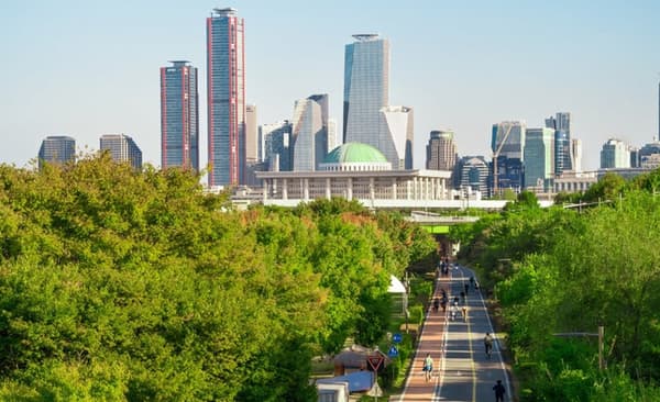 Panoramic view of the Yeouido skyline in Seoul, showing a green park with a bike path and runners in the foreground and tall modern skyscrapers behind the National Assembly building.